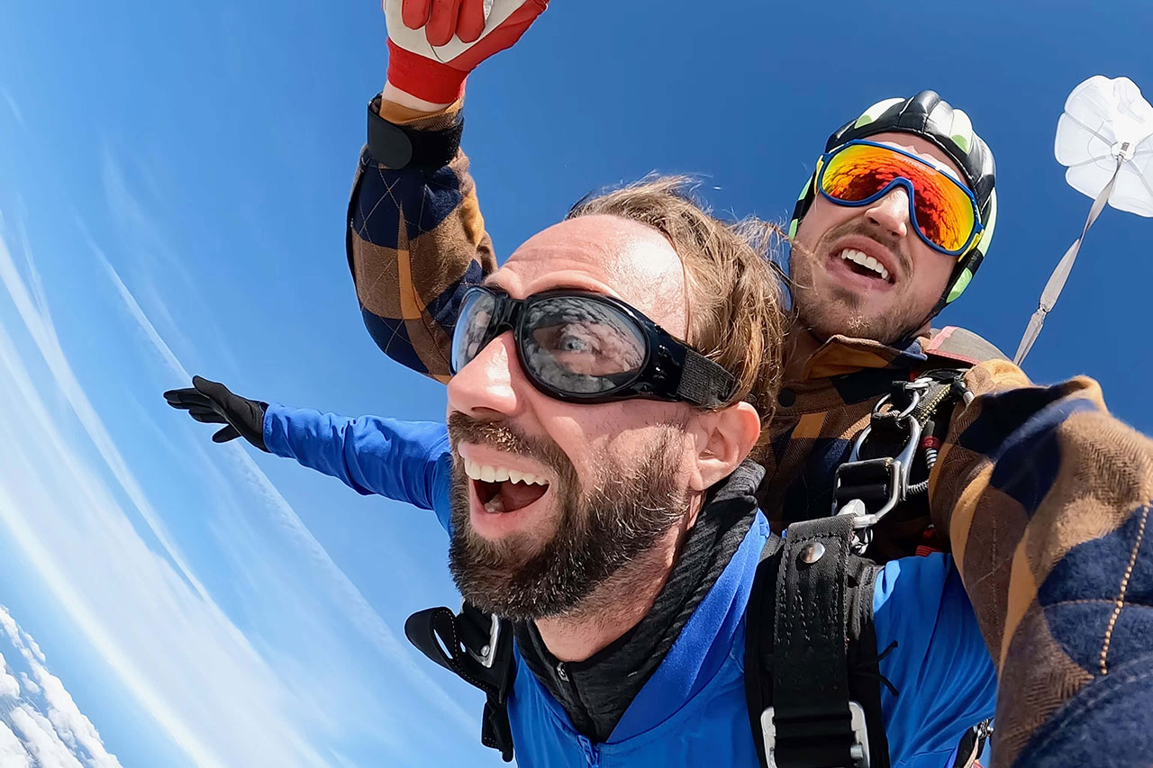 Male tandem pair with goggles and a blue sky and white drogue in the background smiling.