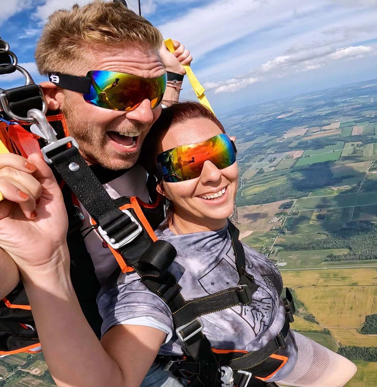 Male and female tandem skydiving pair with rainbow reflective goggles smiling under canopy.