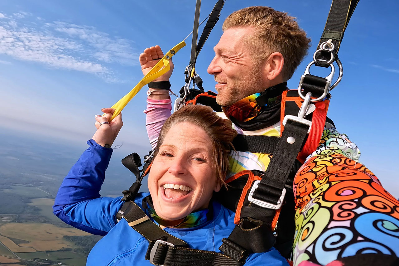 Male tandem instructor in a colorful patterned shirt smiles with female tandem passenger in a blue jumpsuit under canopy.