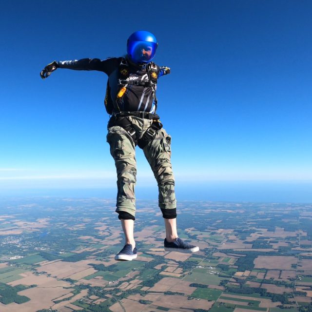 Skydiver in an enclosed blue helmet and camouflage pants flies feet down.