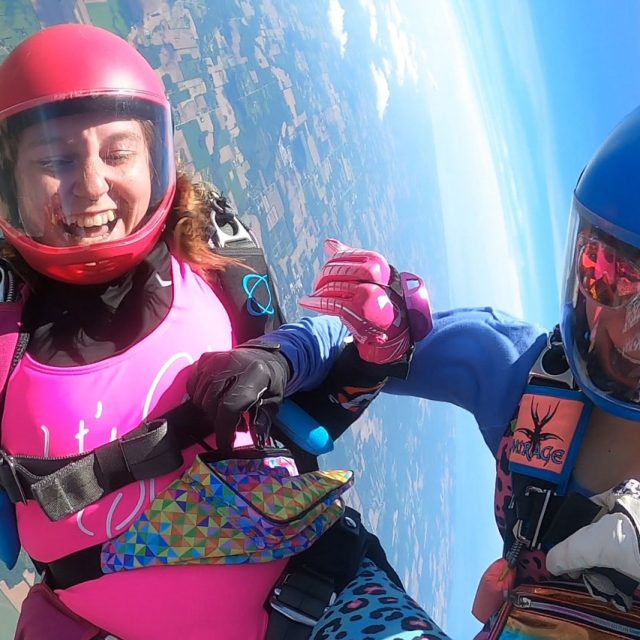 Two female licensed jumpers - one in blue and one in pink- smiling and having a blast in freefall.