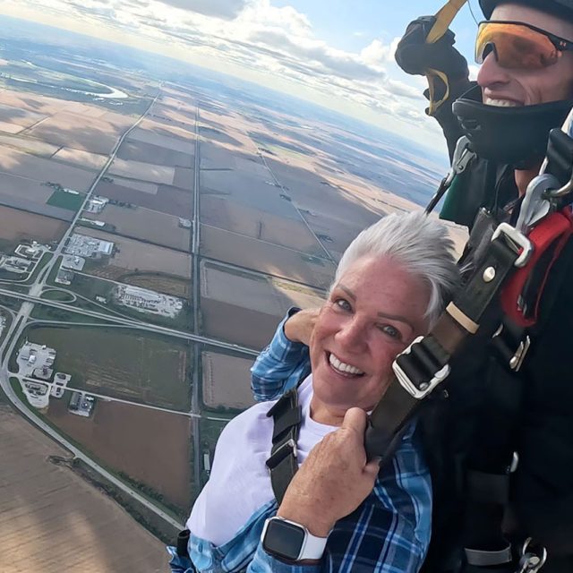 Smiles all around for a male and female tandem skydiving pair under canopy. Farmer fields a-plenty below.