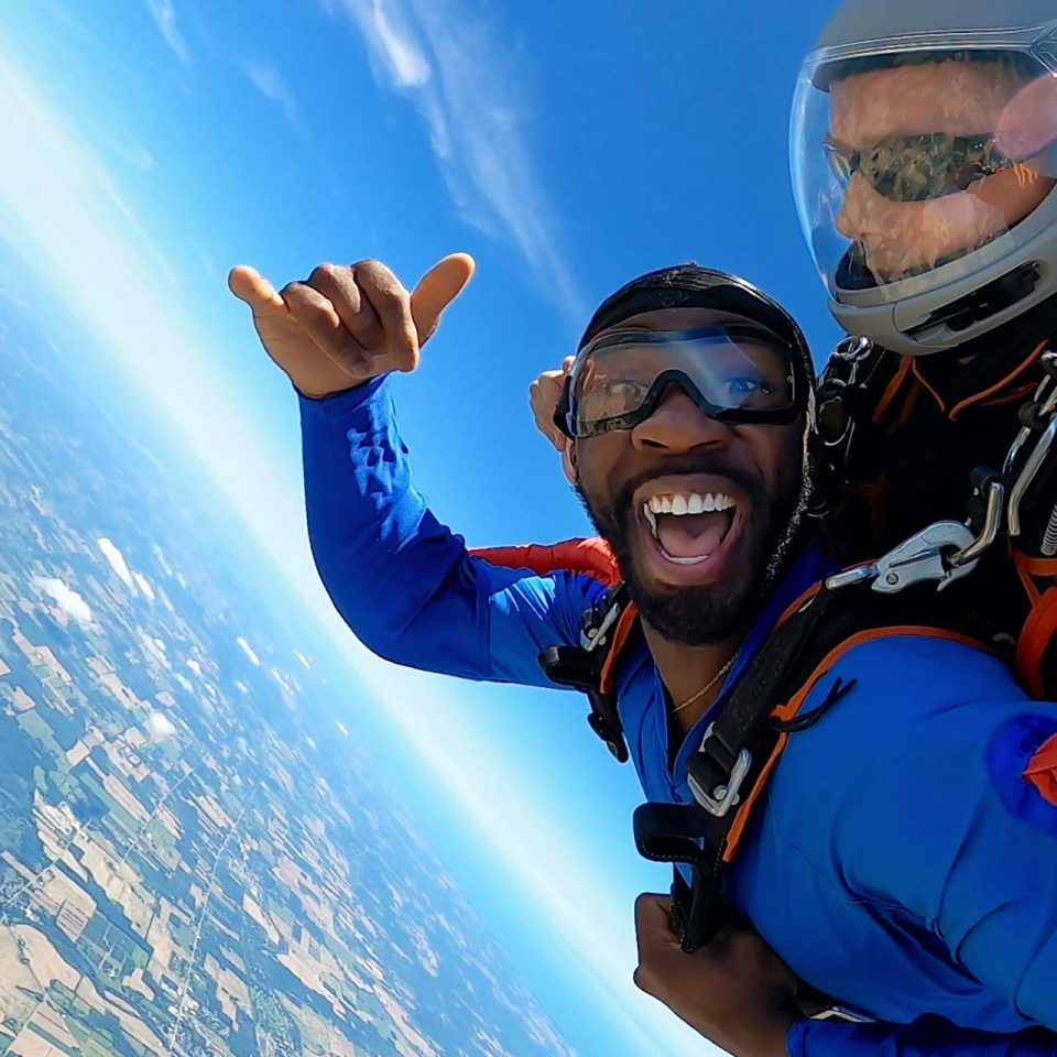 Stoked male tandem skydiving student wearing a blue jumpsuit giving the shaka symbol to the camera.