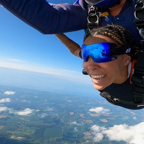 Female tandem skydiving student wearing reflective blue goggles smiles during freefall.