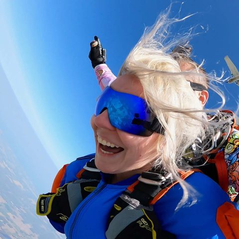 Blonde female tandem student with blue jumpsuit and reflective blue goggles smiles with joy on a tandem skydive.