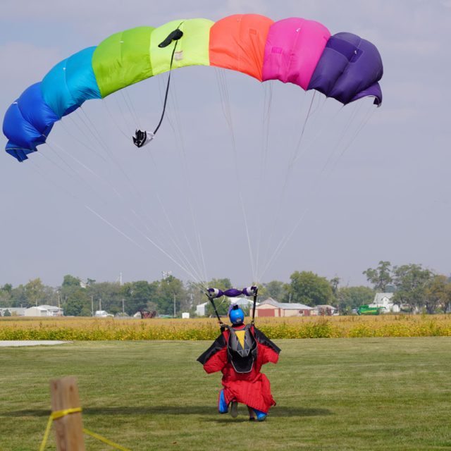 Wingsuiter with a rainbow canopy landing in a green, grassy field.