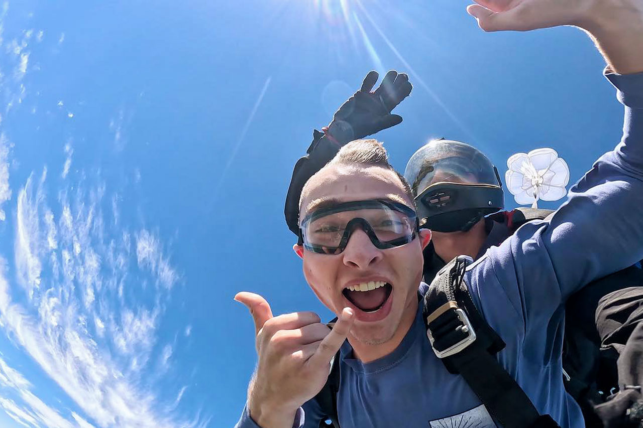 Male tandem skydiving pair giving the shaka hand signal with blue skies and wisps of blue clouds behind.